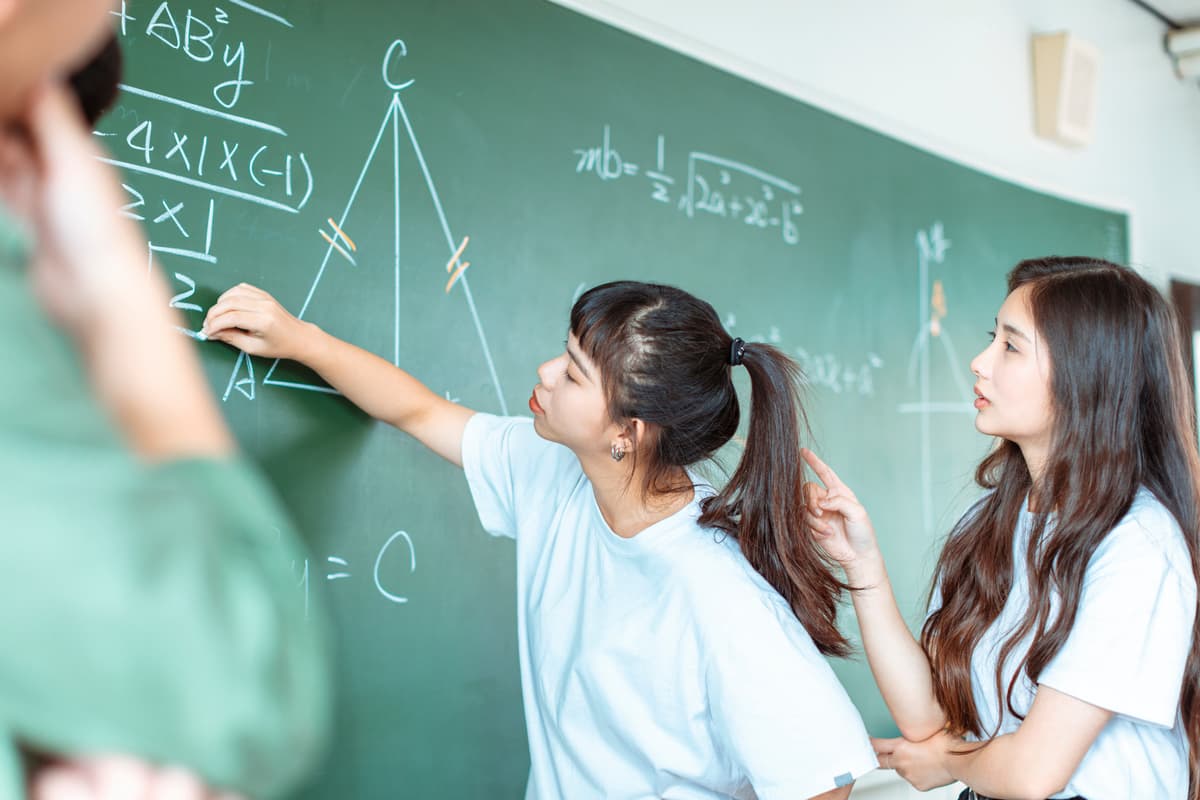 Student solving equations on a chalkboard while classmate observes